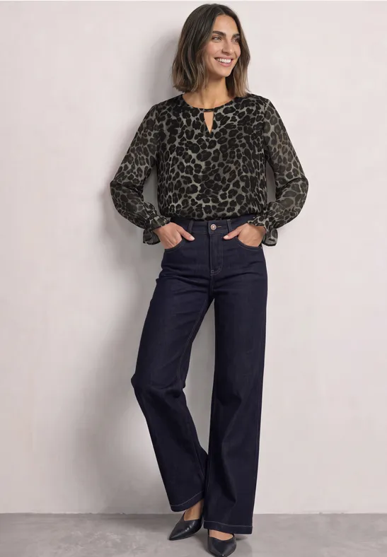 Frau in Leopardenbluse und dunkler Schlagjeans, lächelnd mit Händen in den Taschen.