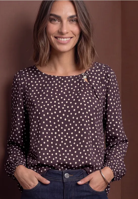 Femme souriante portant un chemisier marron à pois beiges devant un fond marron uni.