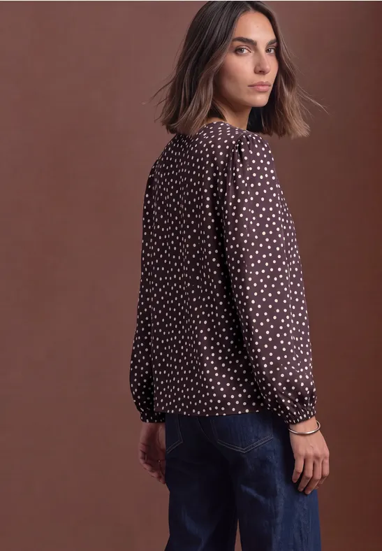 Une femme porte un chemisier marron à pois blancs et un jean, regard par-dessus l'épaule, fond marron.