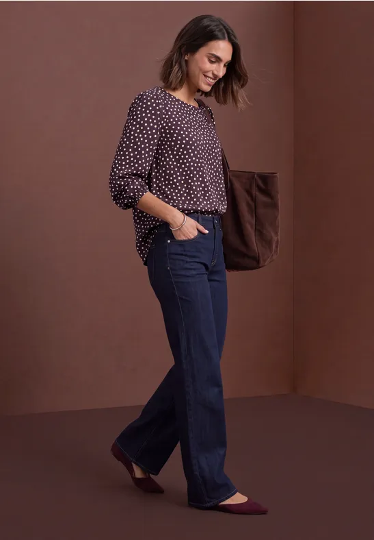 Femme souriante marchant devant un fond marron, vêtue d'un jean ample et d'un chemisier à pois.