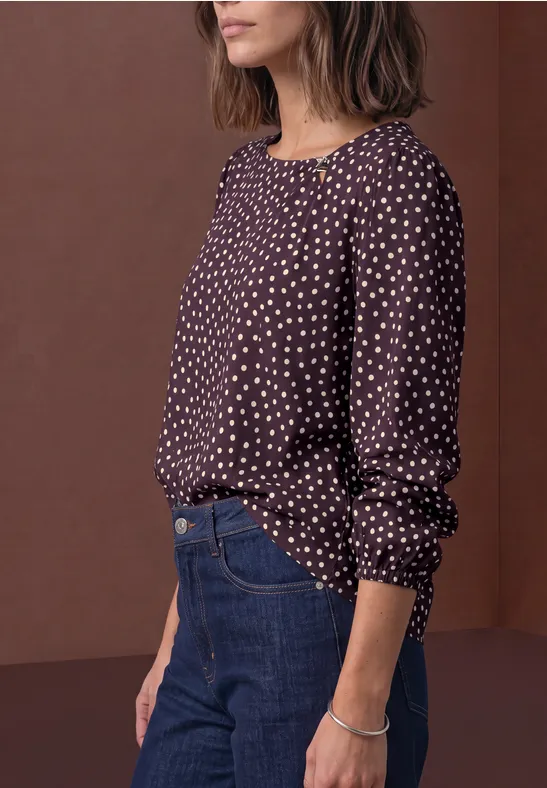 Une femme porte un chemisier foncé à pois blancs, un jean bleu et un bracelet devant un mur marron.