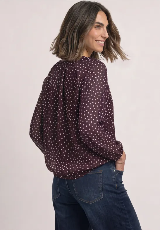 Une femme porte un chemisier bordeaux à pois et un jean, regardant par-dessus son épaule.