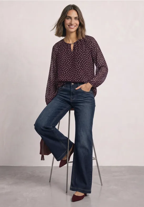 Femme souriante portant un chemisier violet à motifs et un jean évasé, appuyée sur un tabouret haut.