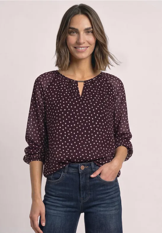 Une femme souriante porte un chemisier bordeaux à pois blancs et un jean foncé, sur fond clair.