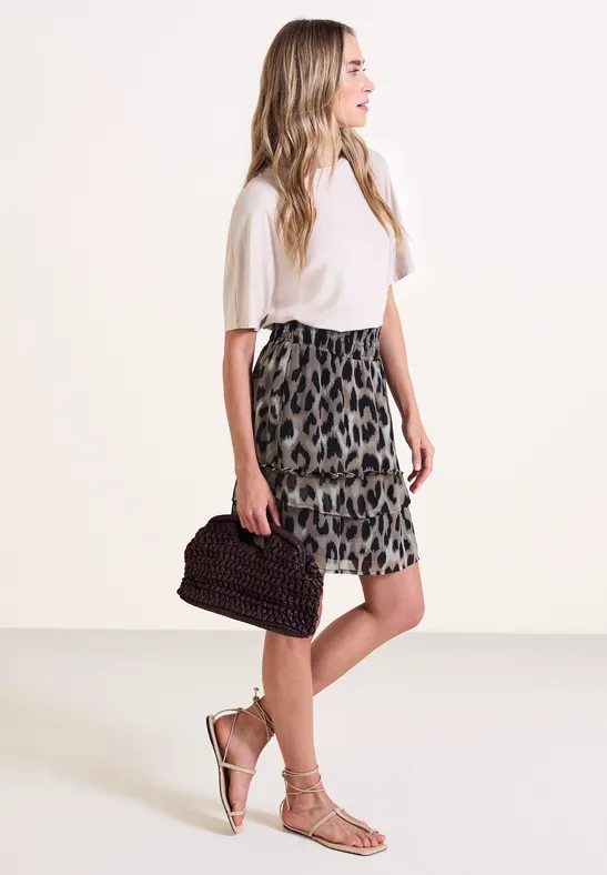 Femme en chemisier blanc, jupe léopard, sandales et pochette, posant de profil sur fond blanc.