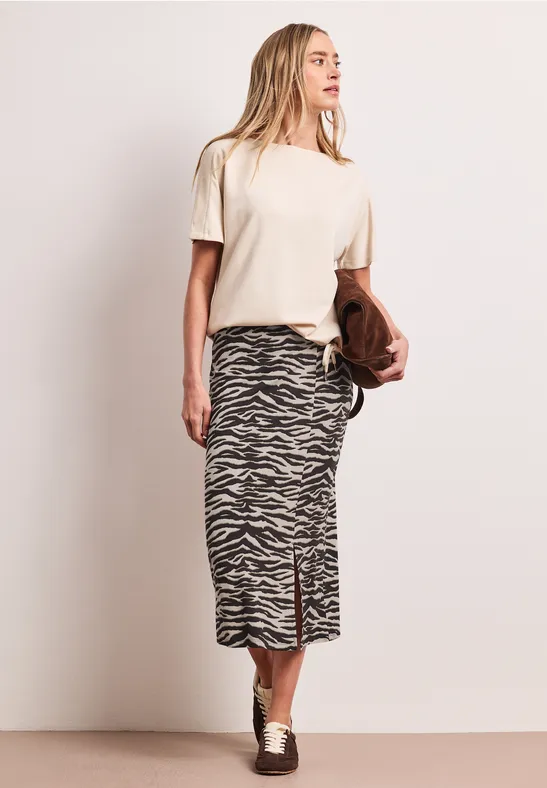 Femme portant un t-shirt blanc, une jupe midi à motif zèbre, des chaussures argentées et un sac marron.
