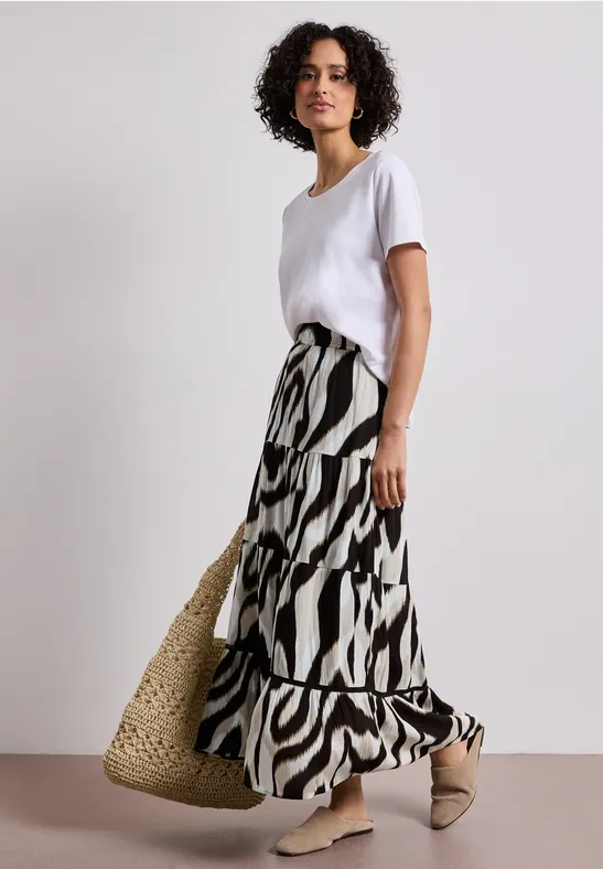 Vrouw in wit T-shirt, lange zebradessin-rok, strooien tas en beige mules voor neutrale achtergrond.