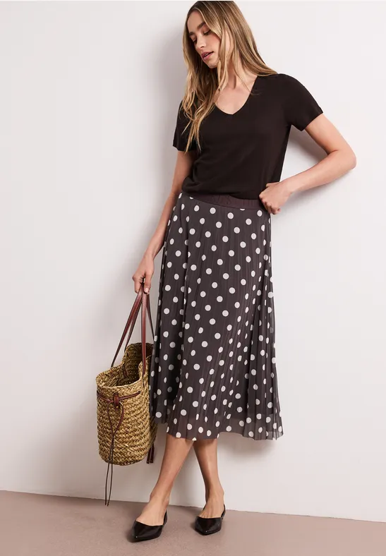 Frau in schwarzem T-Shirt, schwarzem gepunkteten Midirock, Strohtasche und schwarzen Mules.