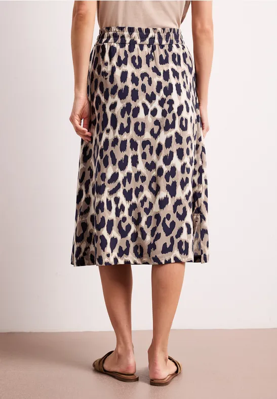 Rückenansicht einer Person in einem knielangen Rock im Leopardenmuster und Sandalen vor neutralem Hintergrund.