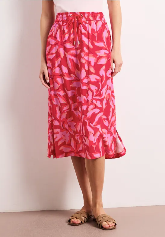 Roter Midirock mit pinkem Blattmuster, locker fallend, kombiniert mit flachen Sandalen auf hellem Untergrund.