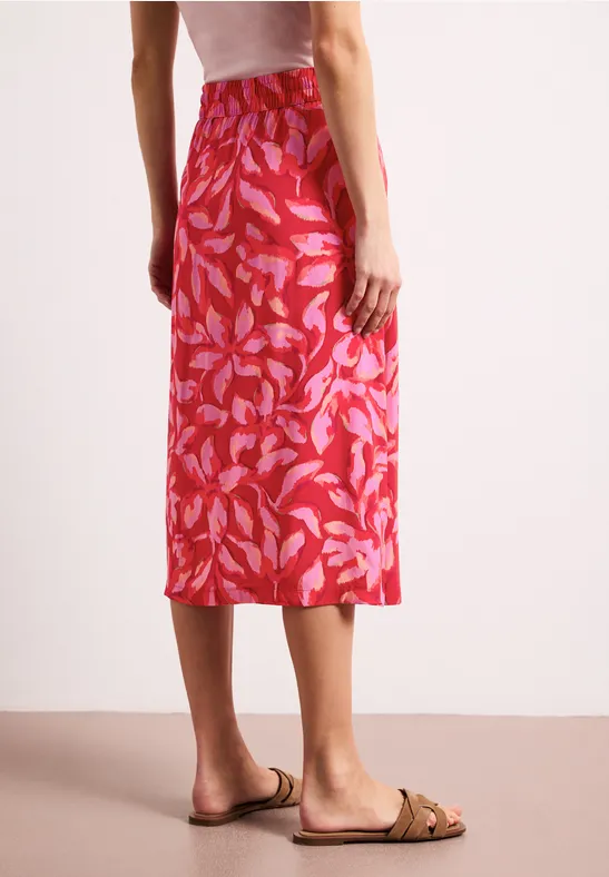 Rückansicht einer Frau in rotem Midirock mit Blattmuster und beigen Sandalen.