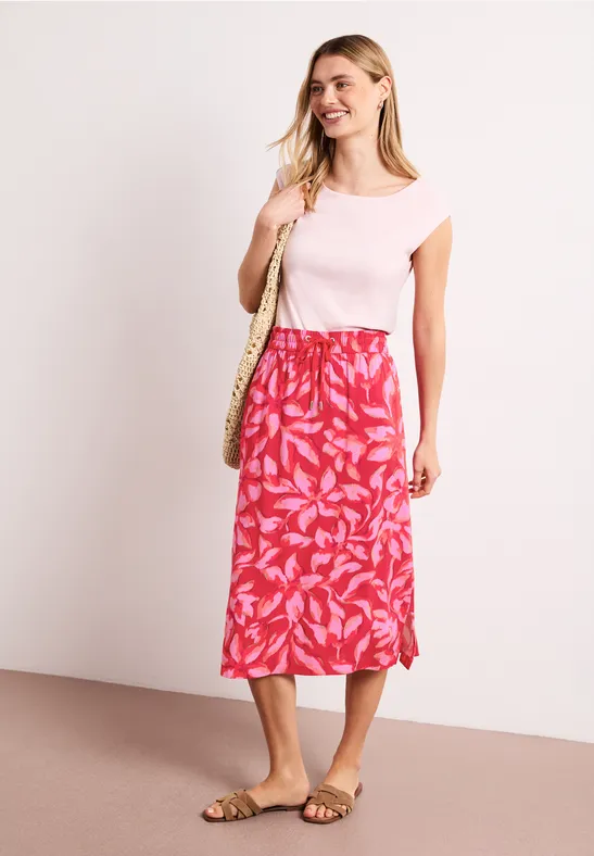 Eine Frau in rosa T-Shirt, rotem Rock mit Blattmuster, Sandalen und Korbtasche vor hellem Hintergrund.