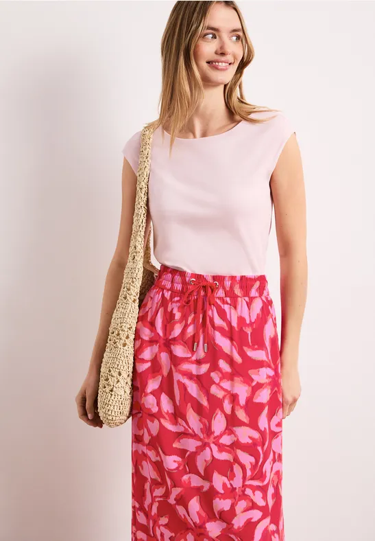 Frau in ärmellosem Rosa-Top, rotem, gemustertem Rock und gehäkelter Schultertasche vor hellem Hintergrund.