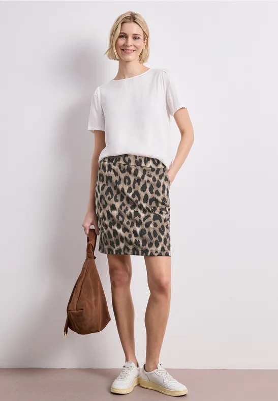 Femme en t-shirt blanc, jupe courte léopard, baskets blanches, porte un sac marron.