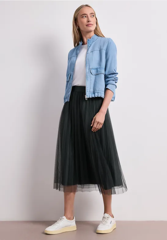 Eine Frau trägt ein schwarzes Tüll-Midirock, weiße Sneaker und eine hellblaue Jeansjacke vor weißem Hintergrund.