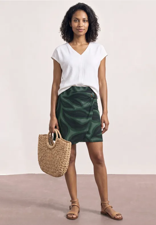 Vrouw met witte blouse, groene rok en sandalen houdt een gevlochten tas voor een lichte achtergrond.