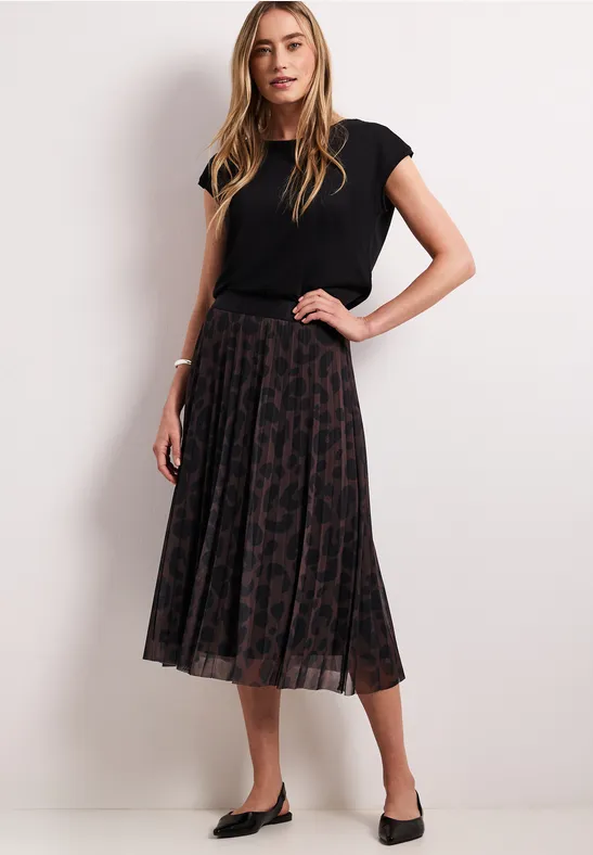 Frau in schwarzem T-Shirt und plissiertem Leo-Midirock mit schwarzen Ballerinas.