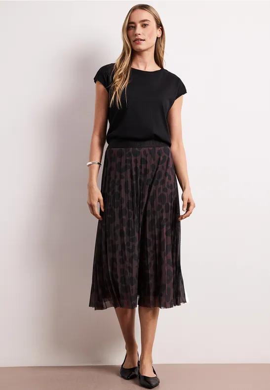 Frau in schwarzem T-Shirt und plissee Midirock mit Muster, dazu flache Ballerinas.