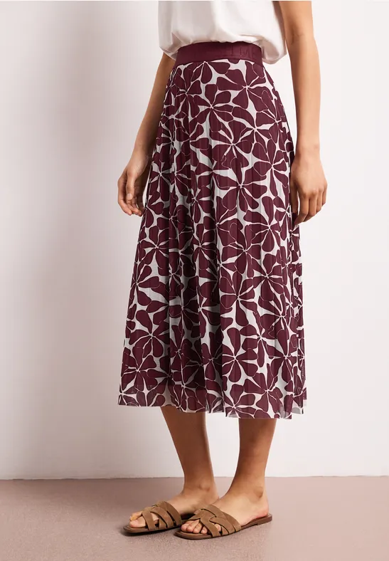 Eine Person trägt einen knielangen lila Rock mit weißem Blumenmuster und flache Sandalen.