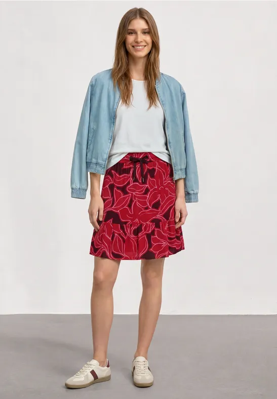 Vrouw met wit topje, rode bloemenrok, lichtblauwe spijkerjack en witte sneakers.
