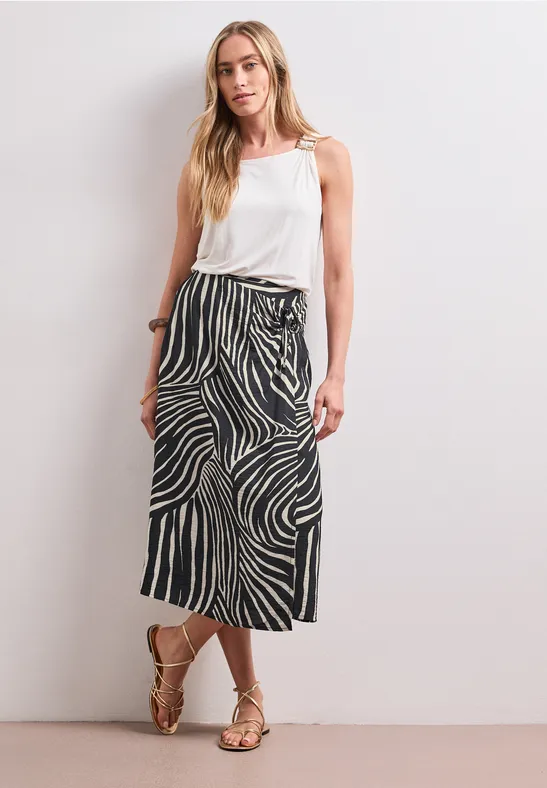 Femme en haut blanc et jupe midi à motifs noirs et blancs devant un mur blanc