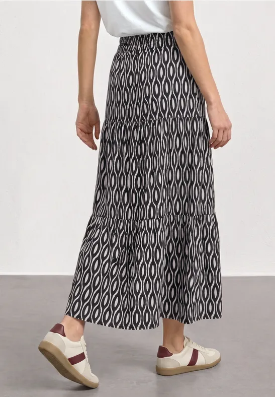 Vrouw in wit T-shirt en lange zwart-witte rok met geometrisch patroon, gezien van achteren, witte sneakers.