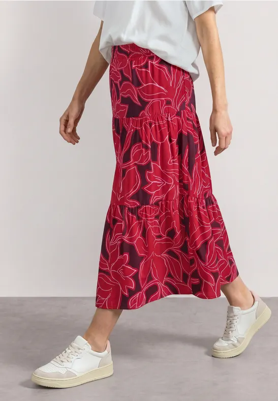 Een persoon draagt een wit t-shirt, een rode midirok met bloemenprint en witte sneakers.