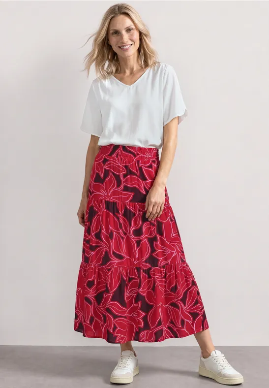 Vrouw in witte t-shirt en lange, rode rok met bladprint, gecombineerd met witte sneakers.