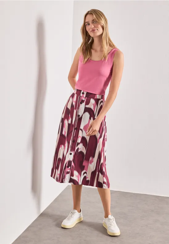 Een vrouw draagt een roze top, gemêleerde midirok en witte sneakers en staat voor een witte muur.