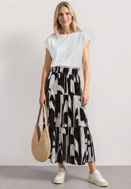 Frau in weißem Top, gemusterten Culottes, weißen Sneakern und Strohtasche vor hellem Hintergrund.