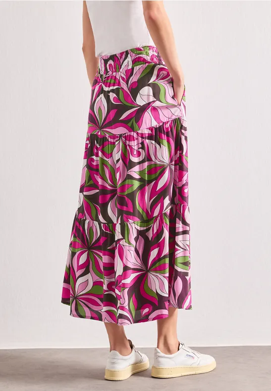 Frau von hinten trägt einen bunten, floralen Maxirock in Pink, Weiß und Grün mit weißen Sneakers.