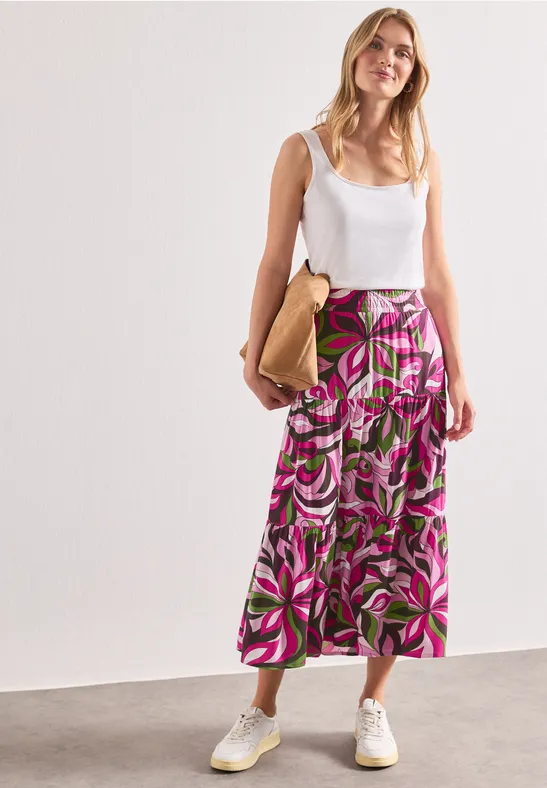Frau in weißem Top, knöchellangem Rock mit pink-grünem Blumenmuster, weißen Sneakern und beiger Tasche.