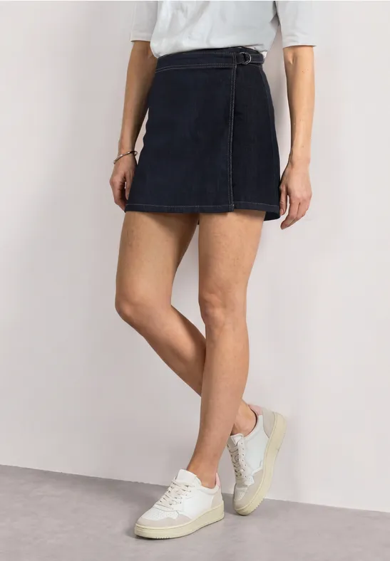 Jupe courte en jean bleu foncé à nouer, sneakers blanches et jambes élancées.