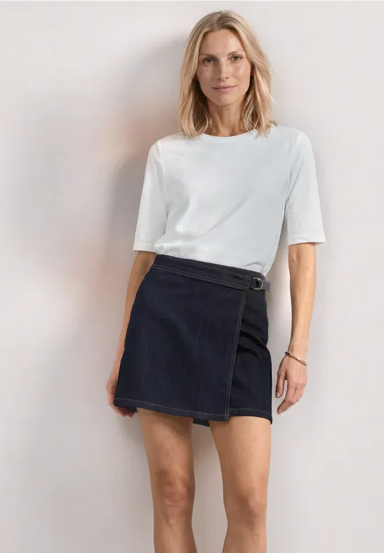 Modèle porte un T-shirt blanc et une mini-jupe en jean foncé sur fond clair.