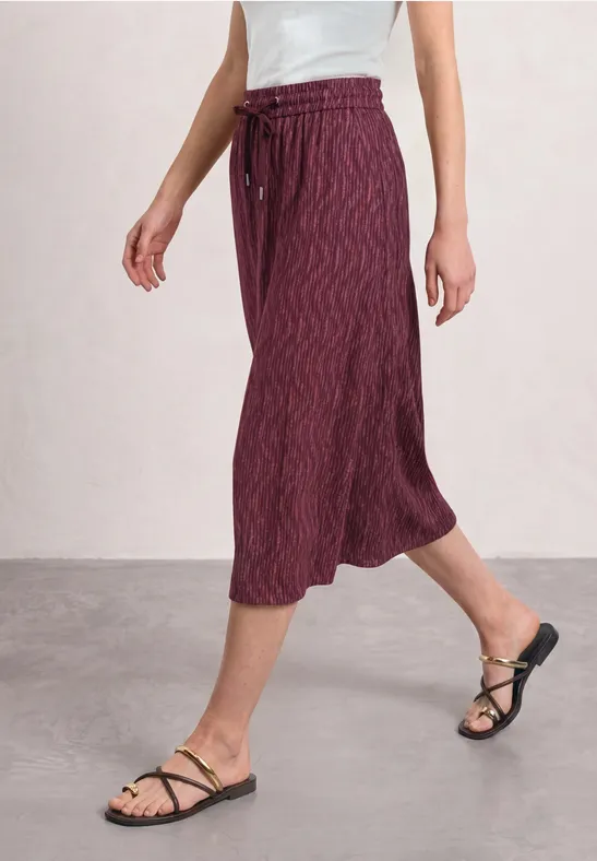 Femme portant une jupe midi bordeaux à motif ondulé et des sandales à brides noires.