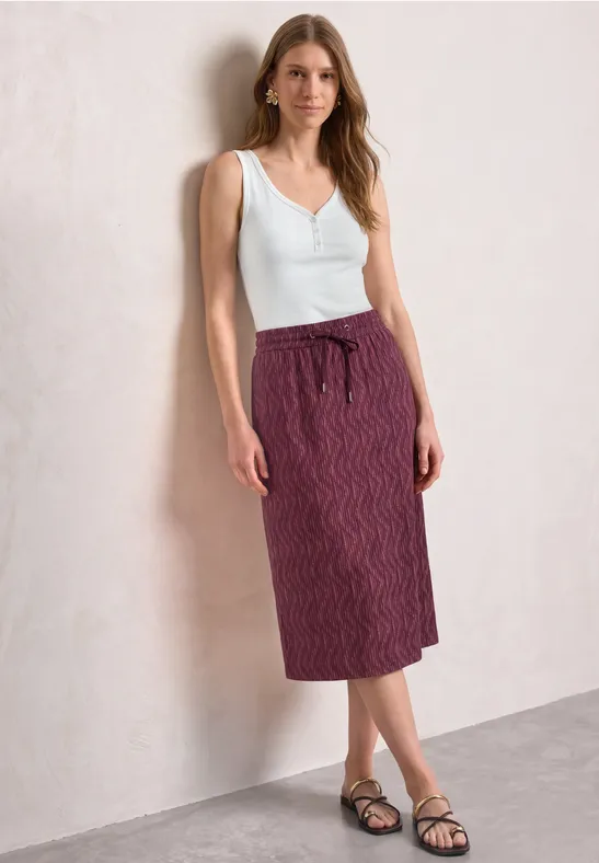 Femme en débardeur blanc, jupe midi violette à motifs et sandales noires, mur clair en arrière-plan.