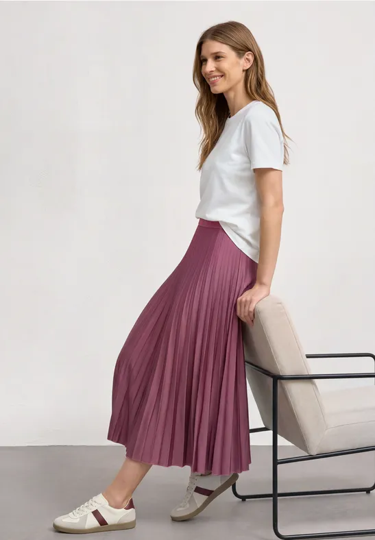 Een vrouw draagt een wit t-shirt en een roze geplooide rok, zittend in profiel op een stoel.