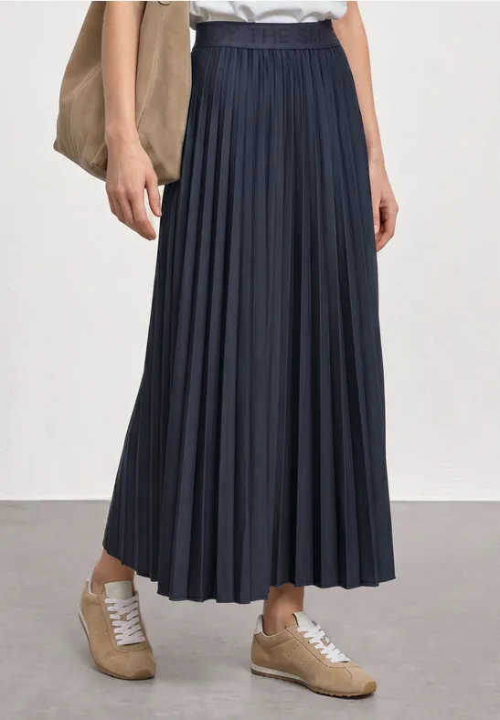 Person trägt dunkelblauen plissierten Midirock, weiße Bluse, beige Sneaker und eine Umhängetasche.
