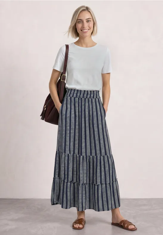 Frau in weißem T-Shirt, gestreiftem Maxirock, braunen Sandalen und Schultertasche vor hellem Hintergrund.