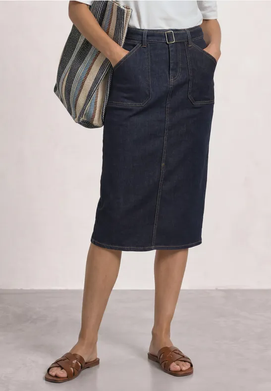 Vrouw met donker denim kokerrok tot de knie, wit shirt, gestreepte tas en bruine sandalen.