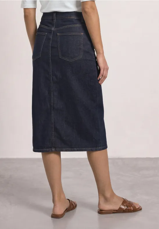 Donkerblauwe jeansmidirok met achtersplit en opgestikte zakken, gecombineerd met bruine platte sandalen.