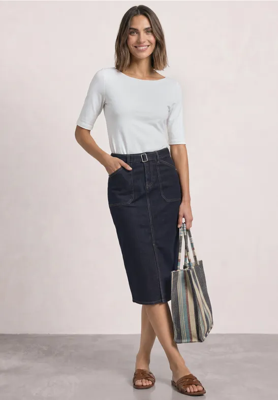 Vrouw in wit shirt, donkere denim midirok en sandalen, met een gestreepte tas.