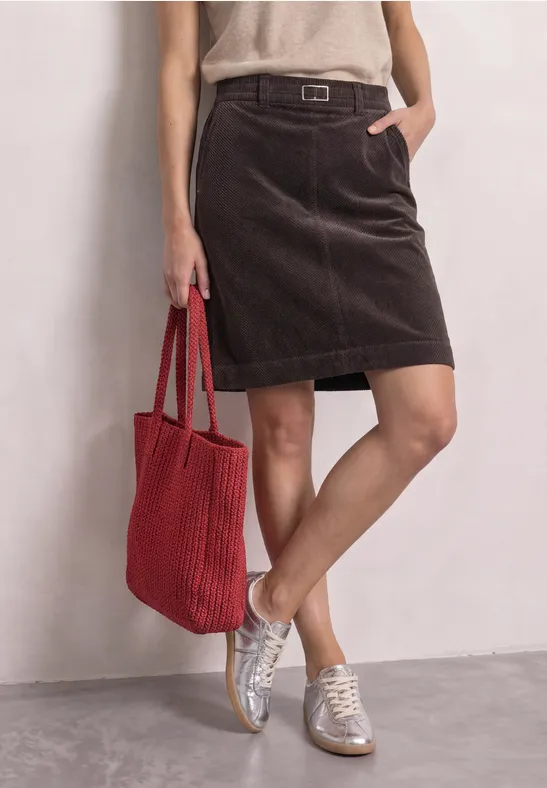 Femme en jupe marron en velours côtelé, top beige, baskets blanches et sac en tissu rouge devant un mur clair.