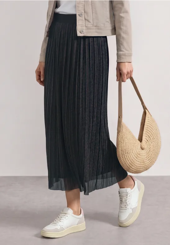 Zwarte plissé midirok, witte sneakers en ronde strooien tas voor een stijlvolle, casual uitstraling.