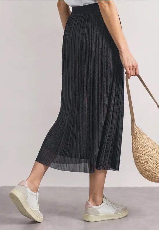 Vrouw draagt een zwarte geplooide midirok, witte sneakers en een gevlochten strooien tas.