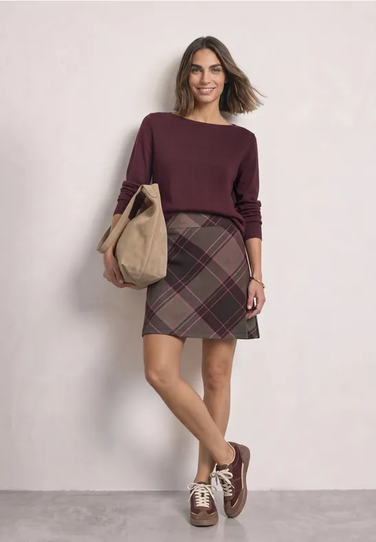Frau in weinrotem Pullover und kariertem Minirock, mit beiger Stofftasche und braunen Sneakers.