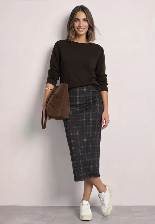 Frau trägt dunkelbraunen Pullover, karrierten Midirock, weiße Sneaker und braune Tasche.