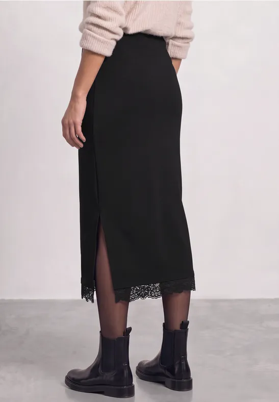 Jupe midi noire fendue sur le côté avec bordure en dentelle, portée avec des boots noires.