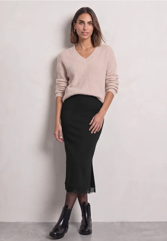 Femme portant un pull beige à col V, une jupe midi noire à dentelle et des bottines devant un mur clair.