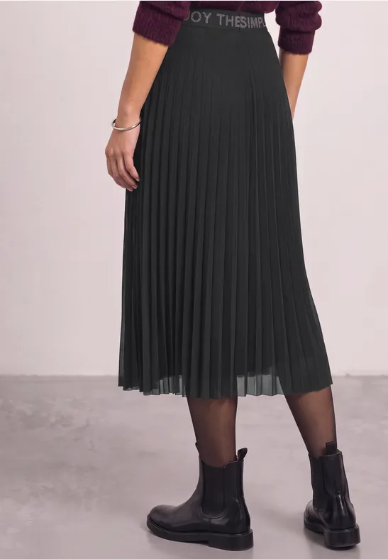 Seitliche Ansicht einer Person in schwarzem, plissiertem Midirock, schwarzen Stiefeletten und Strumpfhose.